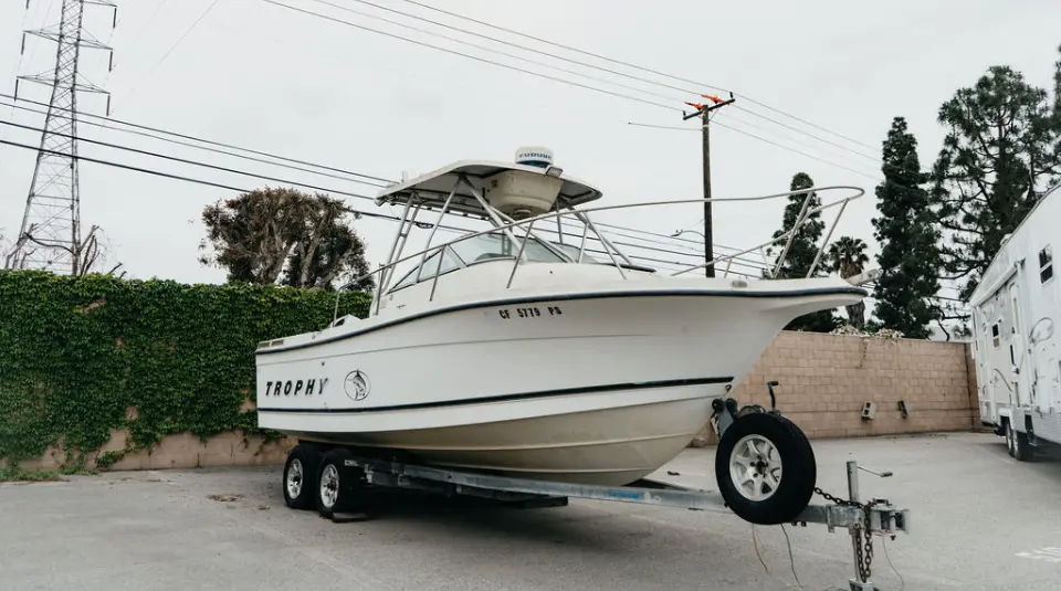 A white Trophy fishing boat on a trailer is parked in a lot next to a wall covered in ivy, with trees and power lines in the background.
