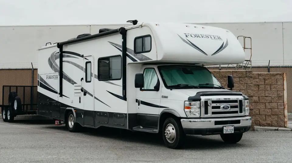 A white Forester motorhome with black and gray accents is parked on a paved lot near a brick wall and industrial building.