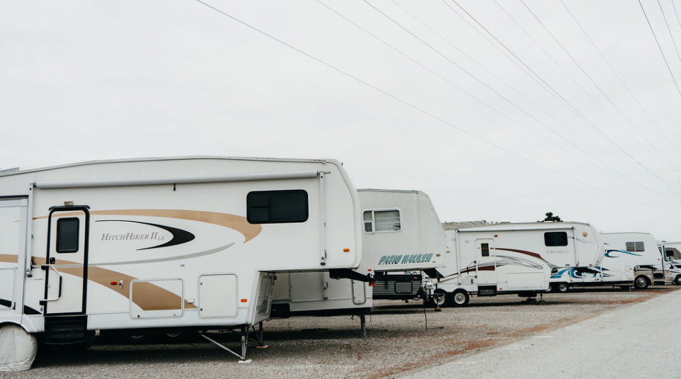 Several RVs and trailers are parked in a row on a gravel lot under an overcast sky, with power lines overhead.