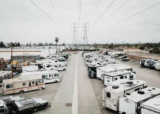 A parking lot filled with numerous RVs and trailers is shown under overcast skies, with power lines and utility poles running overhead.